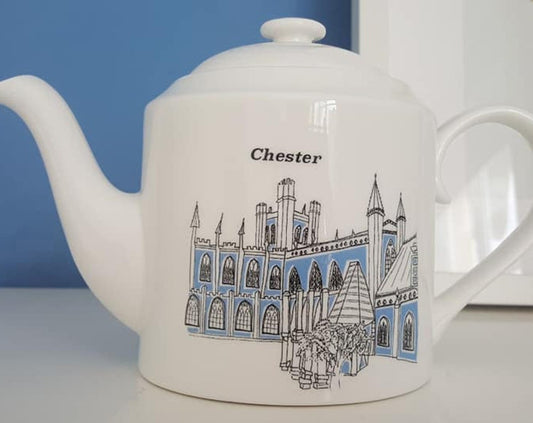 A white ceramic teapot with a blue and black illustration of Chester Cathedral and other buildings on the side, with the word 'Chester' written on the kettle.